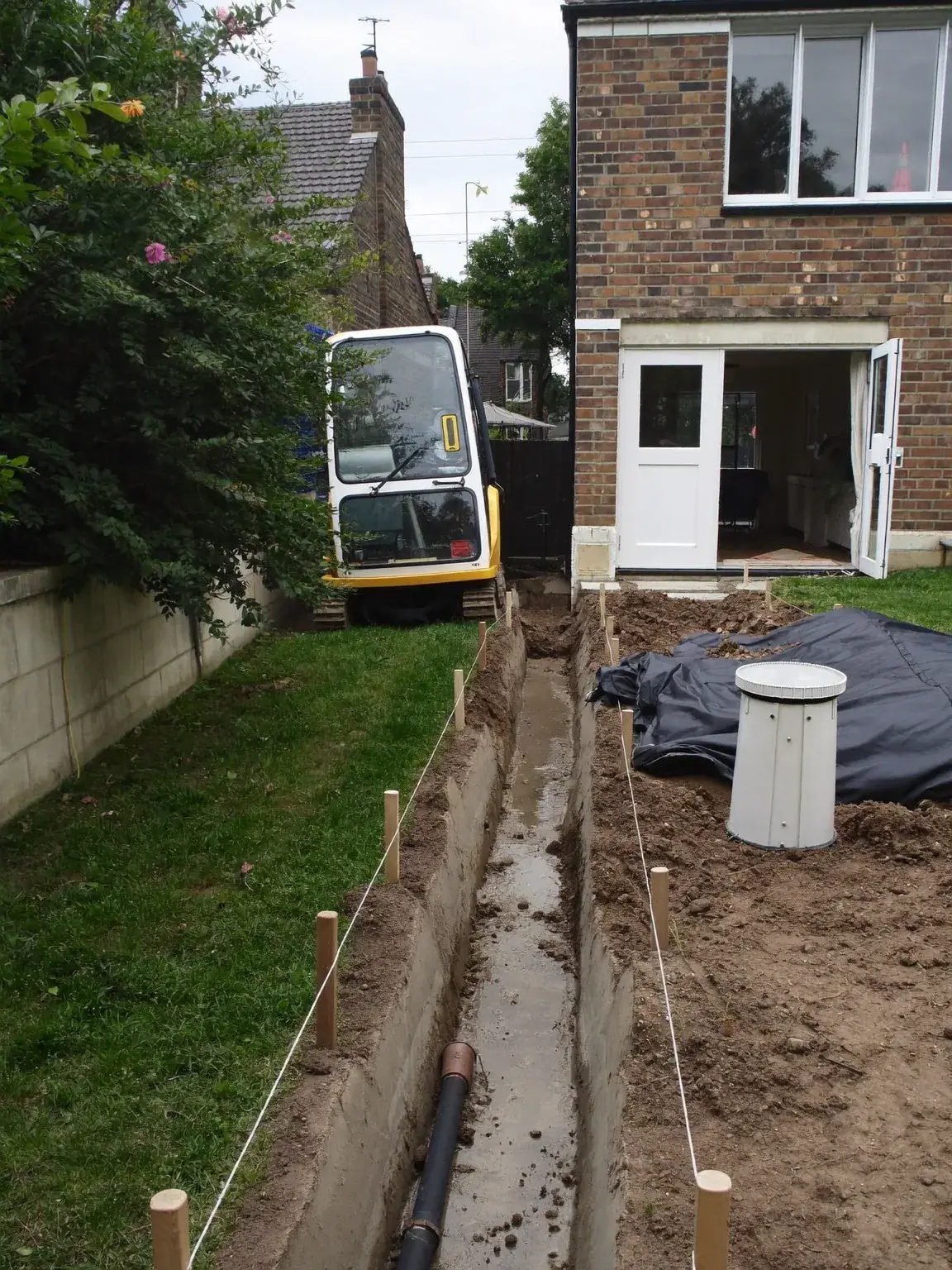 Foundation trenches marked with string lines in a back garden, with a mini digger and a covered inspection chamber