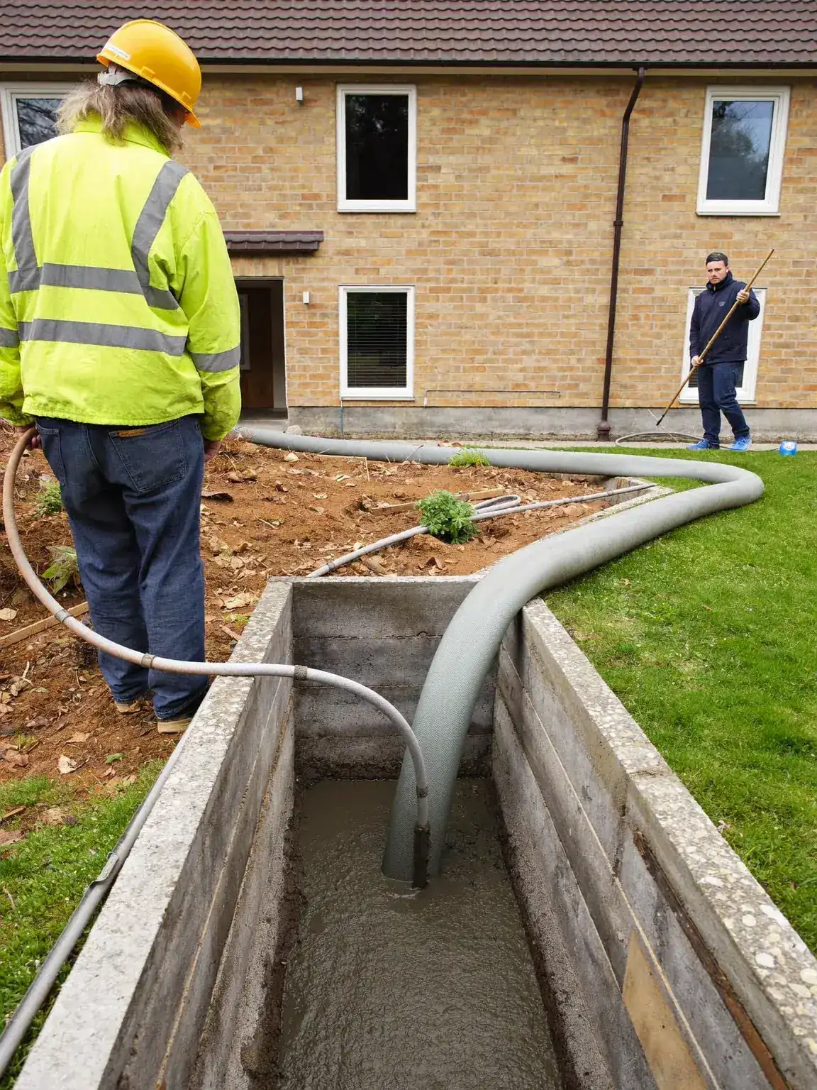 Concrete being poured into a foundation trench through a flexible pump hose, with two workers on site