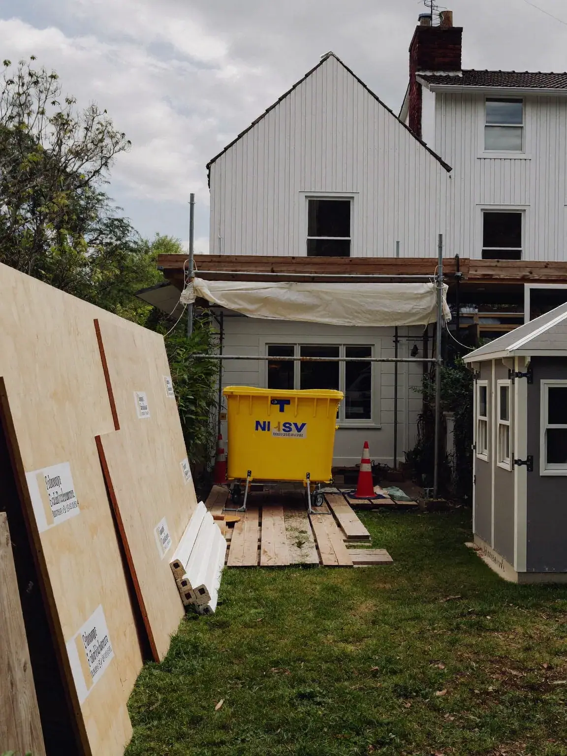 Back garden set up for building work: plywood hoarding, a yellow skip, scaffolding and a garden shed